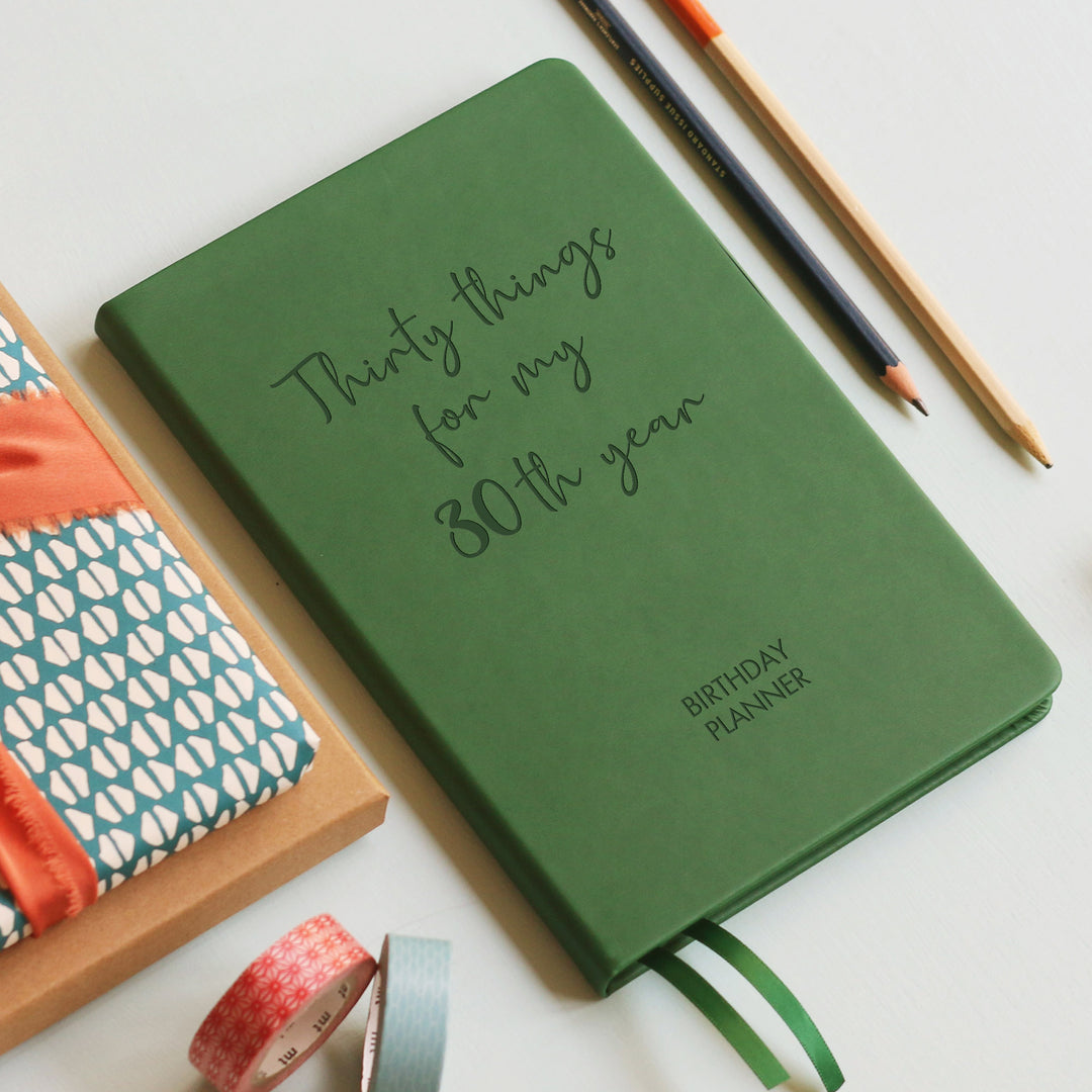 A top down image of a dark green vegan leather notebook resting on a pale aqua table. The front of the notebook features the words thirty things for my 30th year. The personalised engraving reads Birthday Planner. Surrounding the book are various stationary items and dried flowers.