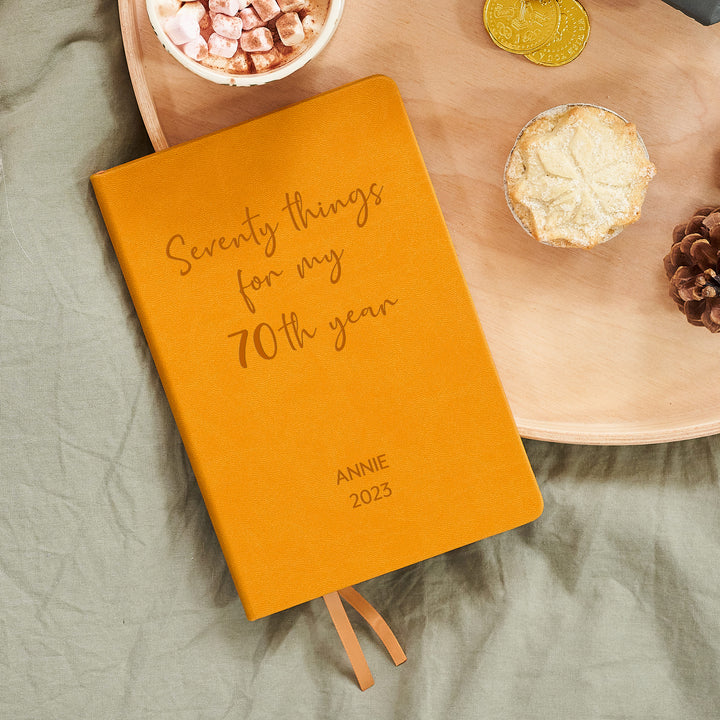 A top down image of a mustard yellow vegan leather notebook. The front of the notebook features the words Seventy things for my 70th year. The personalised engraving reads Annie 2023. It is resting on a wooden tray with a mug of hot chocolate, a mince pie, and a pine cone.