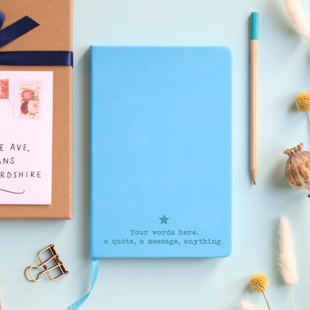 A top down image of a blue hardcover vegan leather notebook resting on a pale aqua table. The bottom of the book is engraved with a personalised message, "Your words here. A quote, a message, anything," and a solid star is positioned above the text. Surrounding the book are various stationary items and dried flowers.