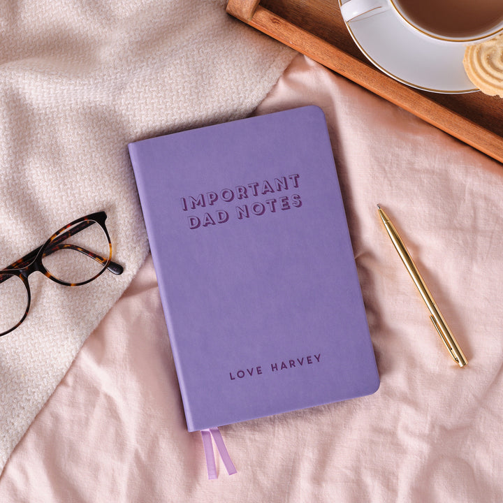 A top down image of a lilac vegan leather notebook resting on a pale pink bed spread. The front of the notebook features personalised engraved text saying IMPORTANT DAD NOTES in large text. LOVE HARVEY is engraved in a smaller text at the bottom of the notebook. Surrounding the book is a tray with a cup of tea and a biscuit, a pair of glasses and a gold pen.