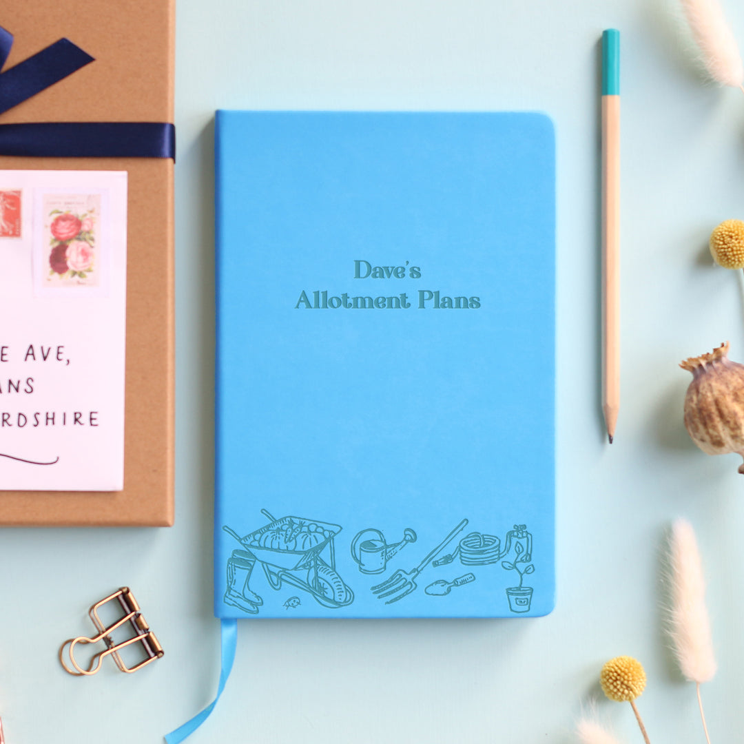 A top down image of a blue vegan leather notebook resting on a pale aqua table. The front of the notebook features an engraving of gardening tools along the bottom edge. The personalised text reads "Dave's Allotment Plans". Surrounding the book are various stationary items and dried flowers.