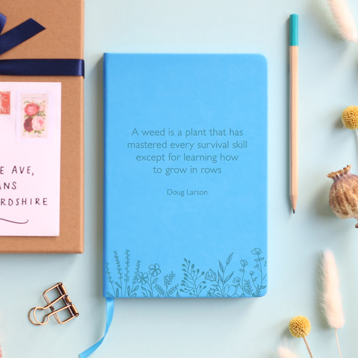 A top down image of a blue vegan leather notebook resting on a pale aqua table. The front of the notebook features a pattern of delicate flowers along the bottom edge. The personalised engraving reads "A weed is a plant that has mastered every survival skill except for learning how to grow in rows - Doug Larson". Surrounding the book are various stationary items and dried flowers.