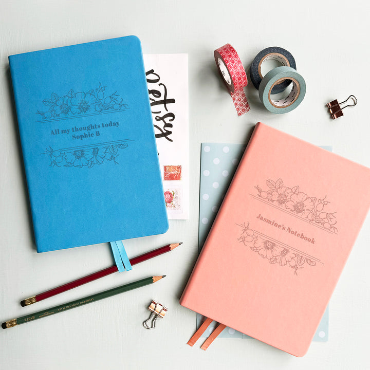 Two notebooks, one pink, one blue, on a pale table with stationery surrounding them. Personalised engraving creates a luxury custom gift!