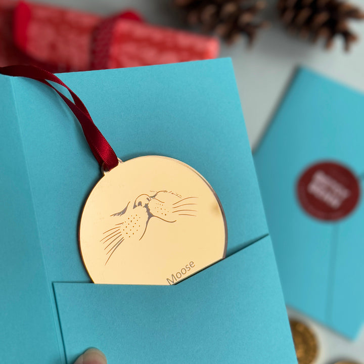Gift presentation blue sleeve holding a gold mirror acrylic Christmas tree decoration with a funny cat face. Shaped like a Gold medal with a red ribbon, with space for a personalised pet name. The perfect custom bauble for a Christmas gift for pet owners, or your Cat sitter!