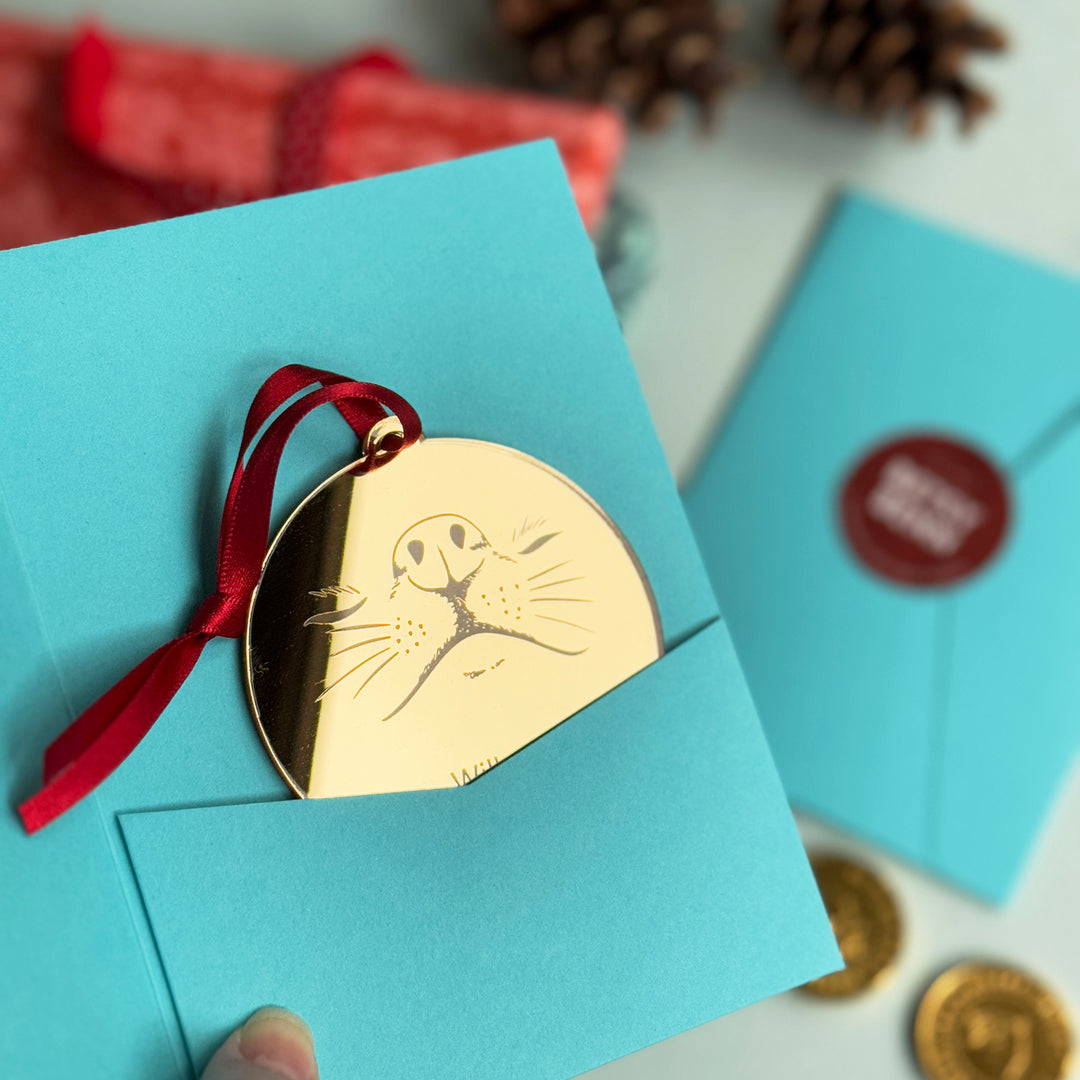 Blue gift sleeve gifting envelope with gold mirror acrylic pet nose dog Christmas decoration and gold coins on a light background, strung with a red ribbon and laser engraved pet memorial or pet sitter gift.