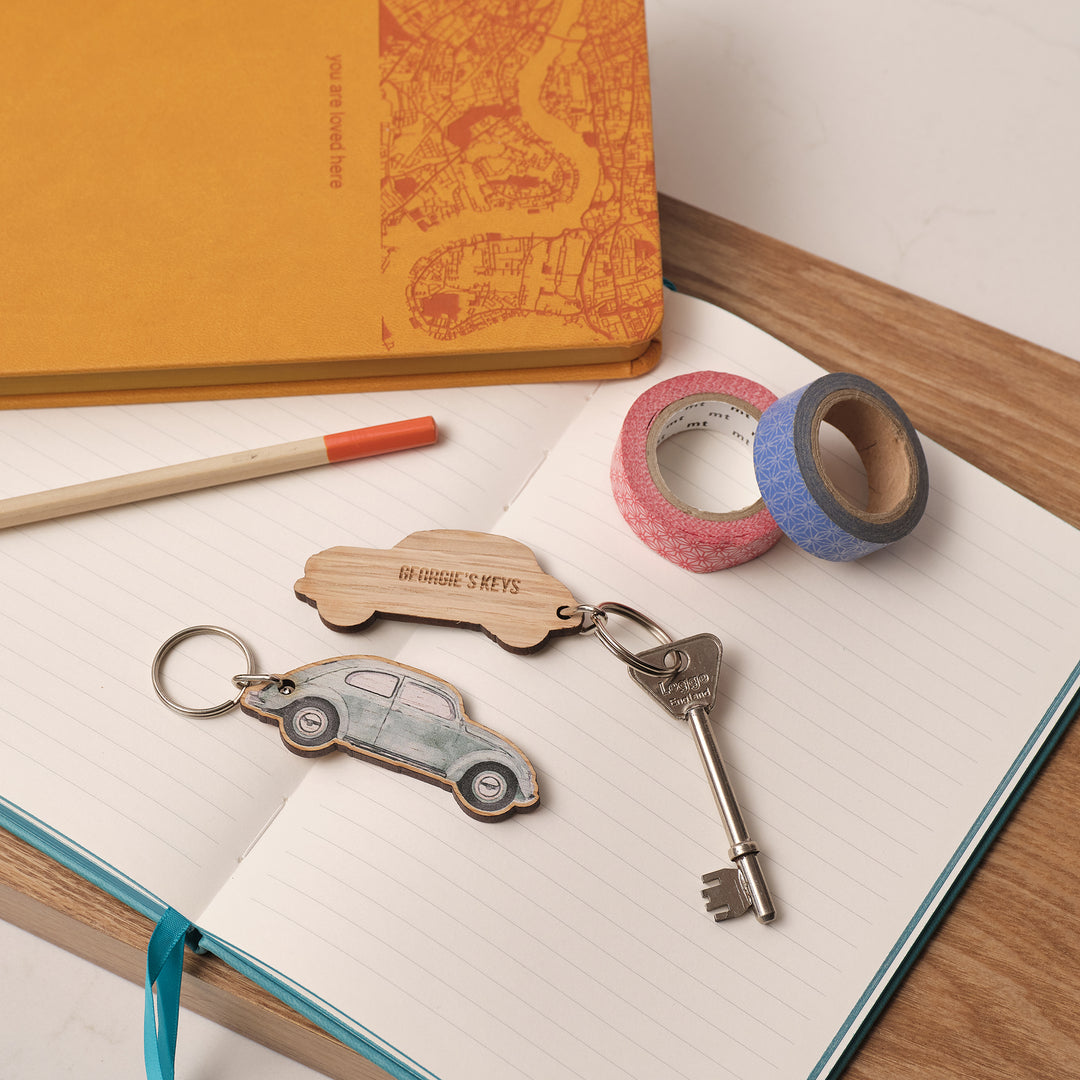 Printed car Volkswagen VW Beetle pale green keychain, key, and decorative tapes on an open notebook with a yellow notebook in the background. Personalise the car gift with a message on the back.