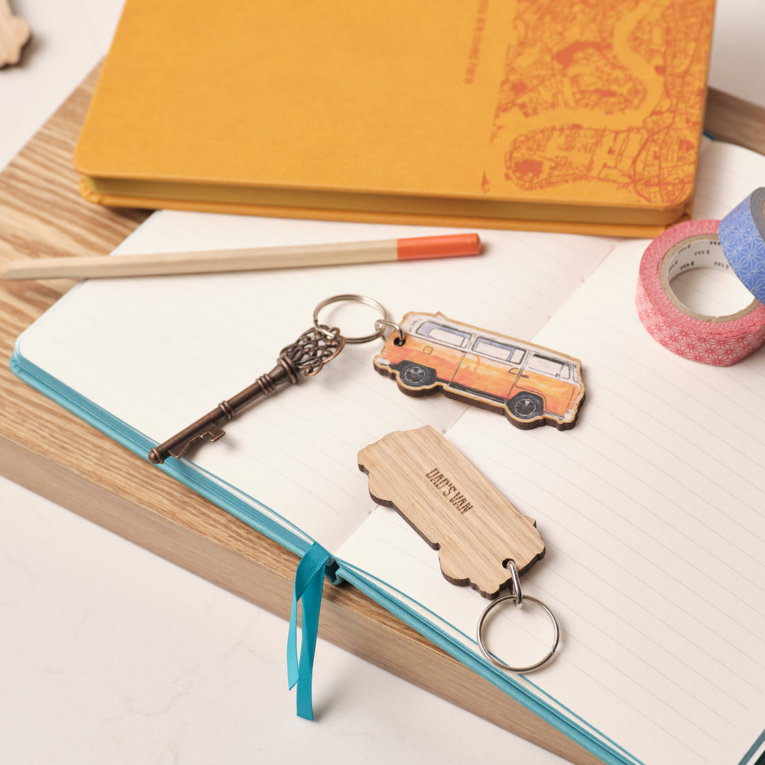 Wooden keychain keyring shaped like an orange VW campervan on an open book with stationery items. Personalised text on the back saying 'Dad's Van'.