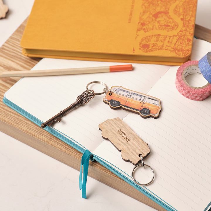 Wooden keychain keyring shaped like an orange VW campervan on an open book with stationery items. Personalised text on the back saying 'Dad's Van'.