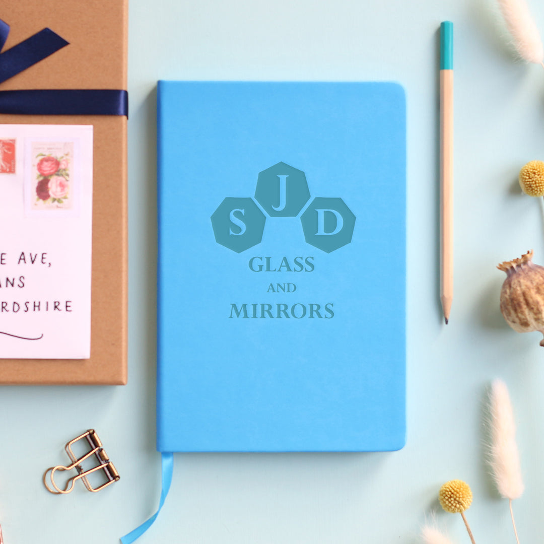 A top down image of a blue vegan leather notebook resting on a pale aqua table. The front of the notebook is personalised with an engraving of a business logo. Surrounding the book are various stationary items and dried flowers.