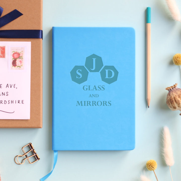 A top down image of a blue vegan leather notebook resting on a pale aqua table. The front of the notebook is personalised with an engraving of a business logo. Surrounding the book are various stationary items and dried flowers.