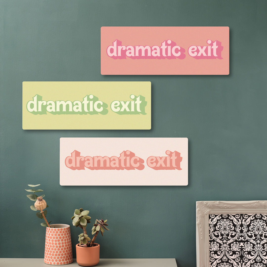 Close-up of a pink and coral wooden sign reading “dramatic exit” in playful retro lettering, mounted on a green wall surrounded by potted plants.