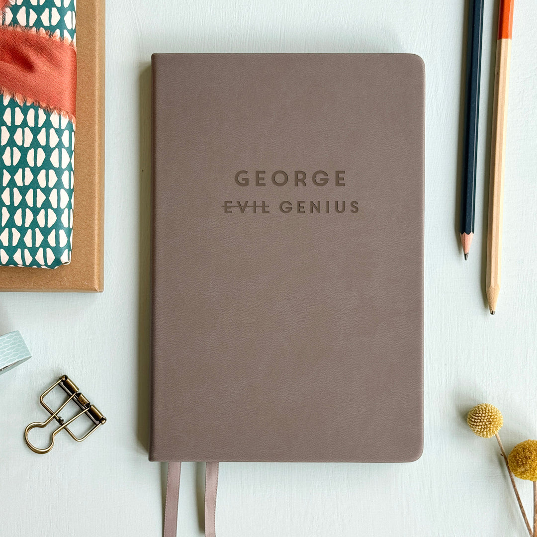 A top down image of a grey vegan leather resting on a pale aqua table. The front of the notebook is personalised with the name George and engraved with the phrase Evil Genius. Surrounding the book are various stationary items.