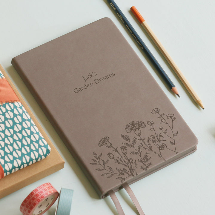 A top down image of a grey vegan leather notebook resting on a pale aqua table. The front of the notebook features a pattern of delicate flowers along the bottom edge. The personalised engraving reads Jack's Garden Dreams. Surrounding the book are various stationary items.