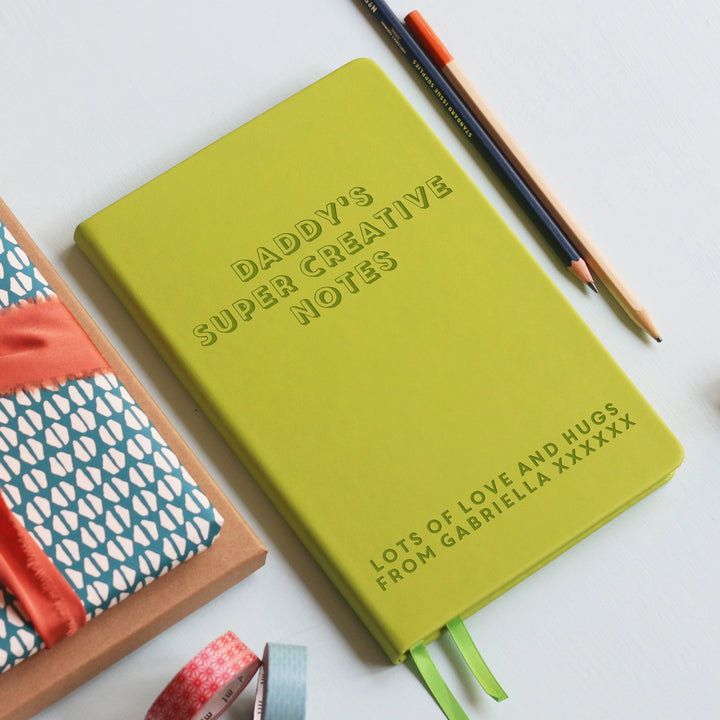 A top down image of a lime green vegan leather notebook resting on a pale aqua table. The front of the notebook is engraved with the personalised text "DADDY'S SUPER CREATIVE NOTES" in a large font. LOTS OF LOVE AND HUGS FROM GABRIELLA XXXXXX  is engraved in a smaller text at the bottom of the notebook. Surrounding the book are various stationary items and dried flowers.