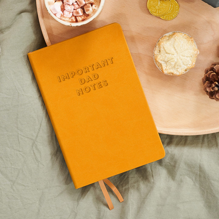 A top down image of a mustard yellow vegan leather notebook. The front of the notebook is engraved with the personalised text "important dad notes" in a large font. It is resting on a wooden tray with a mug of hot chocolate, a mince pie, and a pine cone.