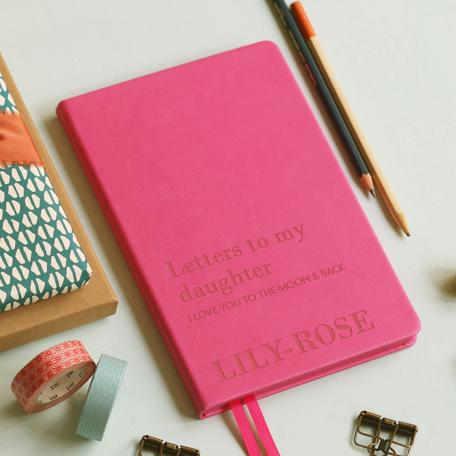 A top down image of a cerise/hot pink vegan leather notebook resting on a pale aqua table. The front of the notebook is engraved with the phrase "Letters to our daughter" in large text and underneath it is personalised with the words I LOVE YOU TO THE MOON AND BACK in a smaller font size. Personalised with the name Lily-Rose. Surrounding the book are various stationary items.
