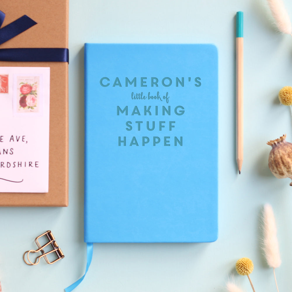 A top down image of a pale blue vegan leather notebook resting on a pale aqua table. The front of the notebook is personalised with the text  “Cameron's little book of making stuff happen". Surrounding the book are various stationary items and dried flowers.