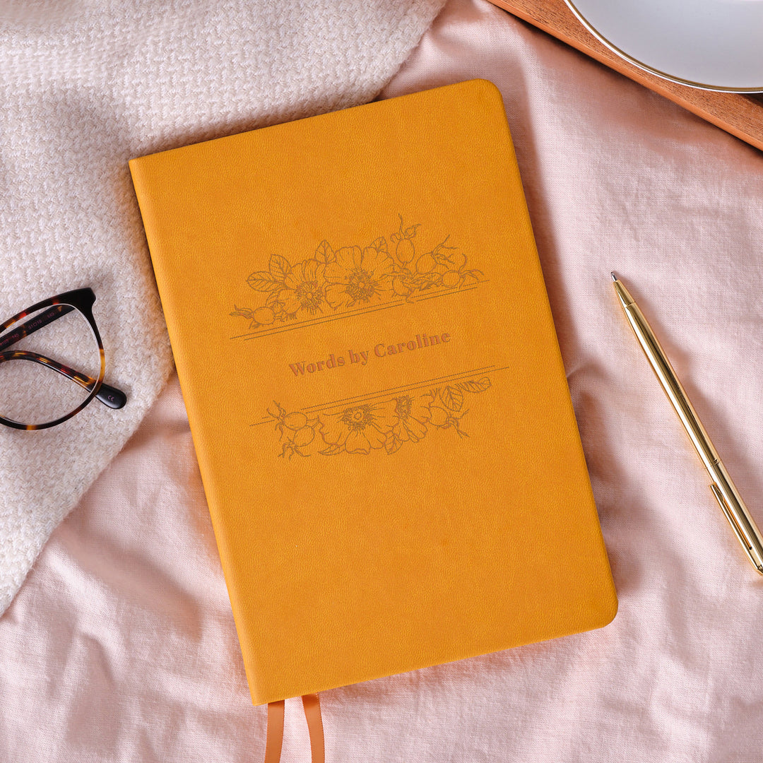Mustard Yellow floral engraved vegan leather A5 lined notebook with colourful ribbons, placed on a pale pink bedspread next to a pen and some glasses.