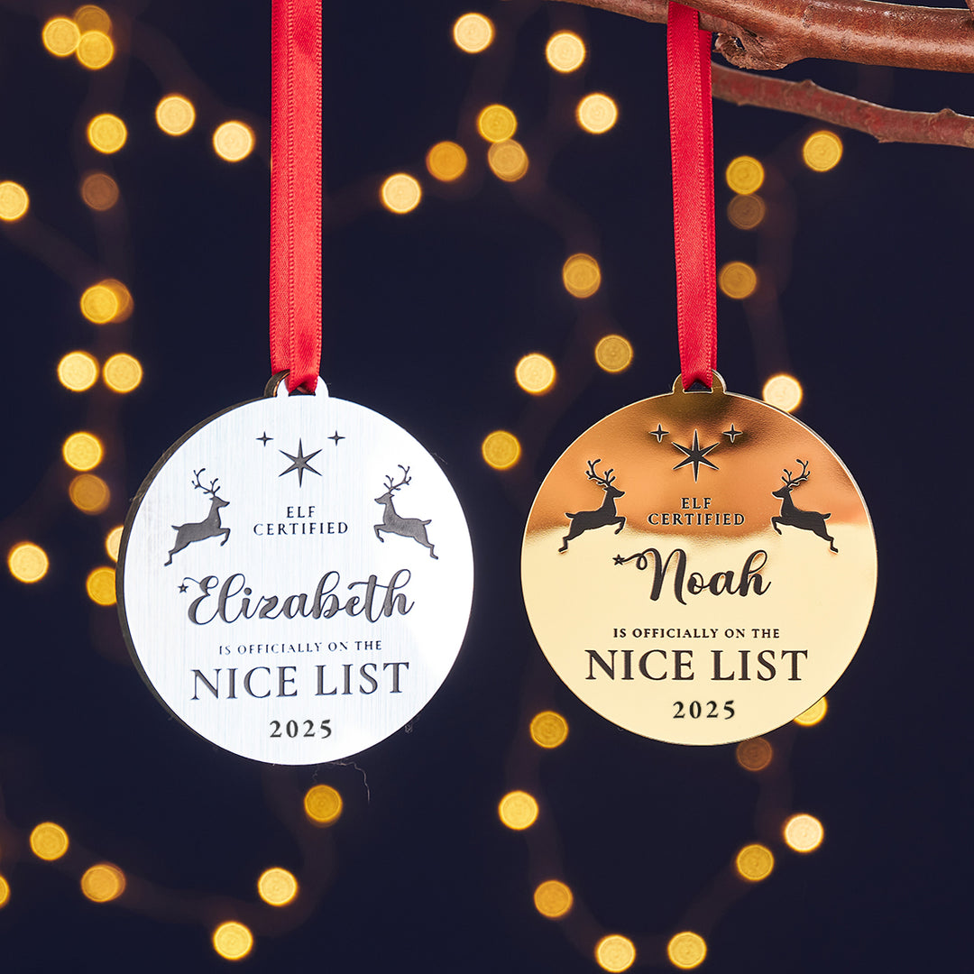 Two circular personalized Christmas tree decorations, one with a silver reindeer and text reading 'Elf Certified - Officially on the Nice List 2025', and the other with a gold reindeer and text 'Nice List 2025'. Both are mounted on red satin ribbons and cedar wood veneer, with a blurred Christmas tree and lights in the background.
