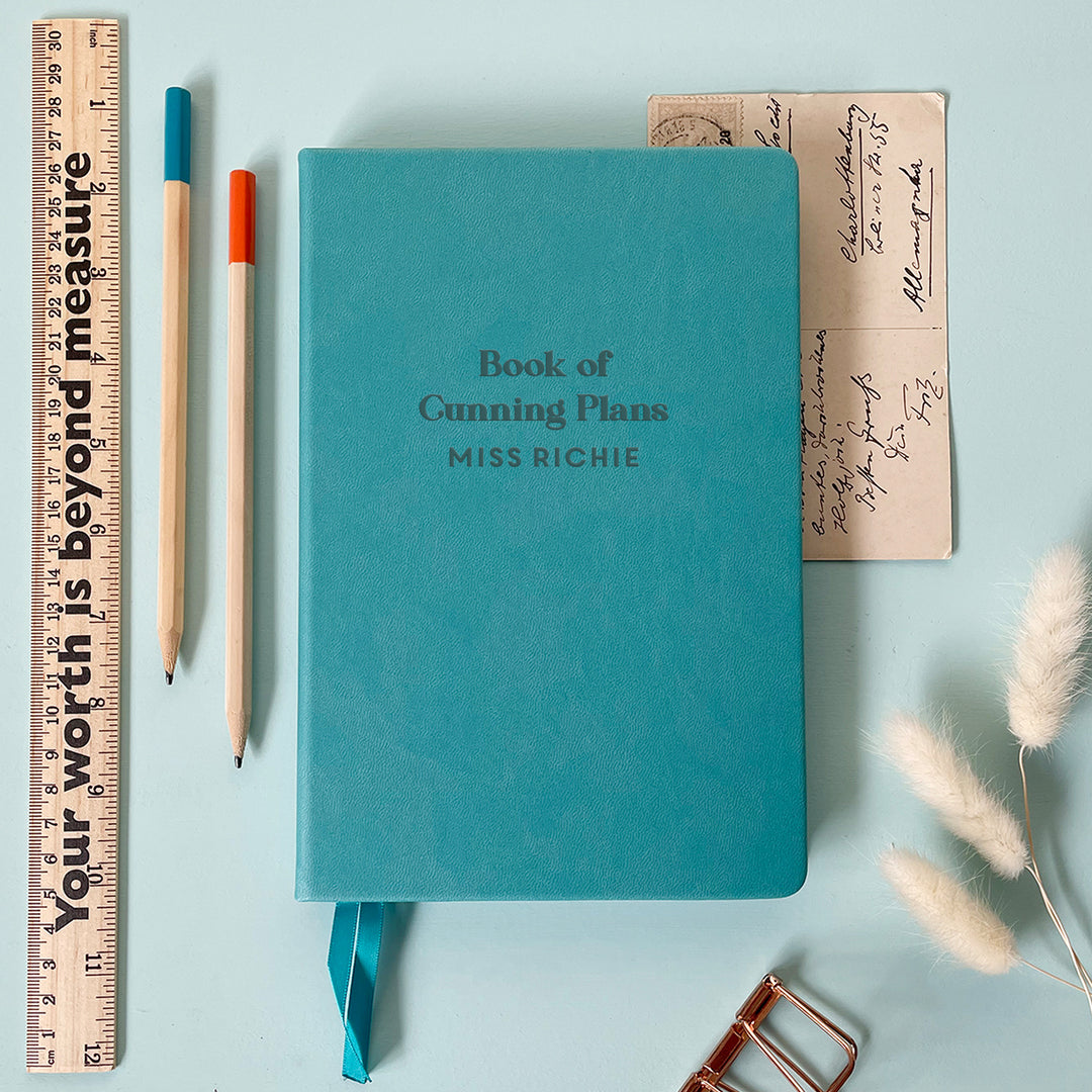 A flat lay image of a turquoise vegan leather notebook resting on a pale aqua table. The front of the notebook is engraved with the phrase “Book of Cunning Plans” and is personalised with the name Miss Richie. Surrounding the book are various stationary items and dried flowers.
