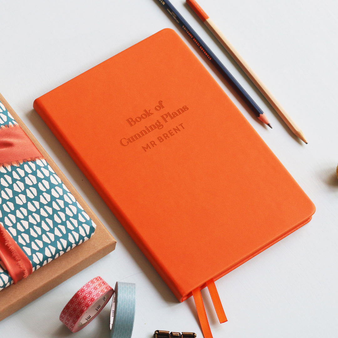 A flat lay image of a orange vegan leather notebook resting on a pale aqua table. The front of the notebook is engraved with the phrase “Book of Cunning Plans” and is personalised with the name Mr Brent. Surrounding the book are various stationary items.
