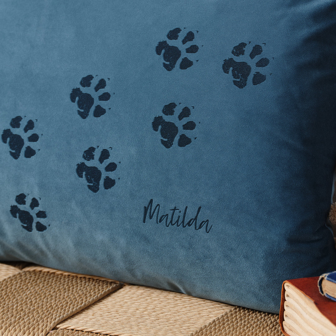 A close-up of a blue velvet cushion with paw prints and the name "Matilda" embroidered on it. The cushion is resting on a woven surface with books nearby.
