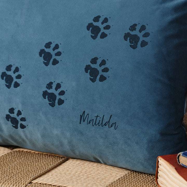 A close-up of a blue velvet cushion with paw prints and the name "Matilda" embroidered on it. The cushion is resting on a woven surface with books nearby.