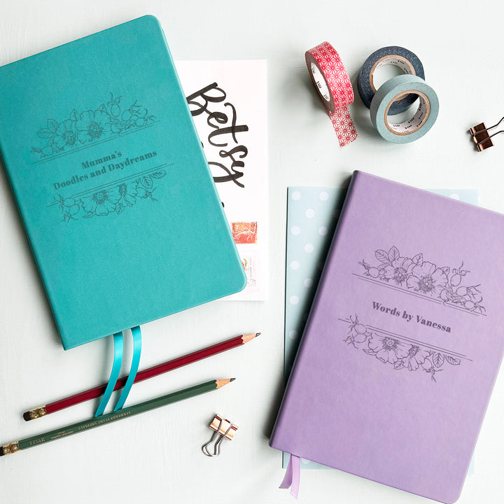 Two floral notebooks in lilac and turquoise with personalised text, perfect gift for notetakers, laid on a white background accompanied by colourful stationery.