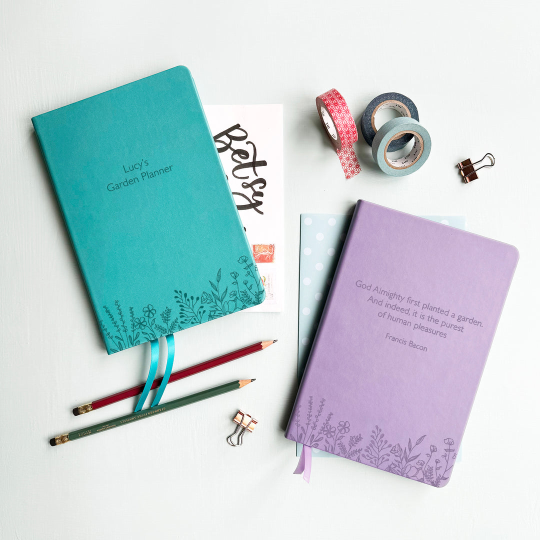 A top down image of two vegan leather notebooks, one turquoise and one lilac. The front of the notebook features a pattern of delicate flowers along the bottom edge. The personalised text reads Lucy's Planner on the turquoise notebook and on the lilac a quote from Francis Bacon. Surrounding the book are various stationary items.