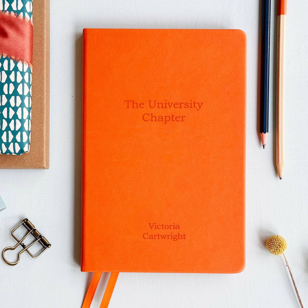 An orange personalized notebook with 'The University Chapter' text on a light blue background with decorative items. Custom back to school graduation gift or teacher school supplies.