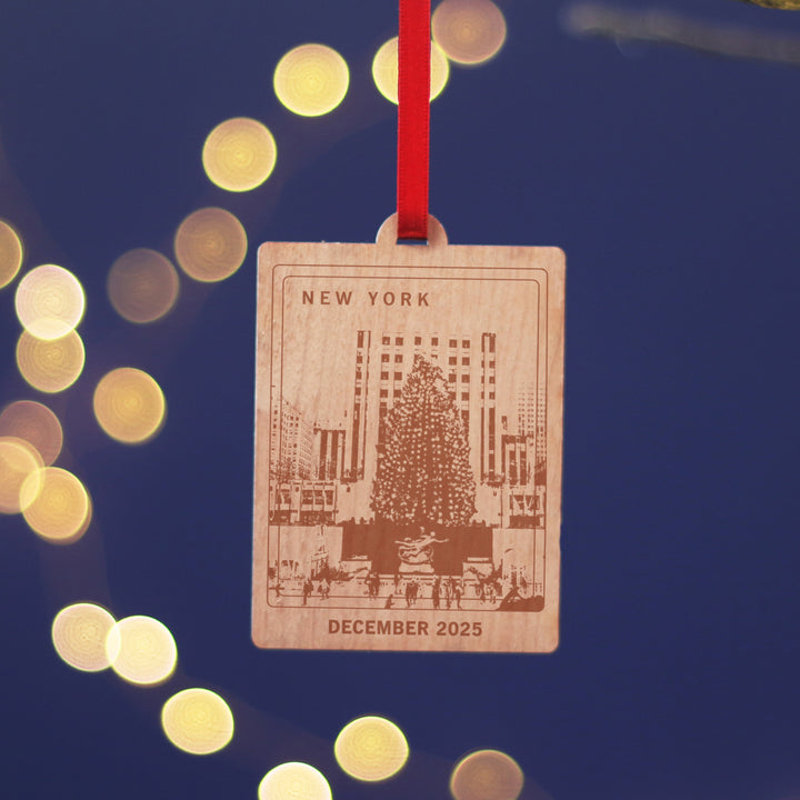 New York travel memory anniversary gift, commemorate your Christmas memories, with this sustainable wood cherry veneered ornament with Rockefeller Center and ice skaters in front of a Christmas tree. Hanging against a blue background. Personalise the location and text for a perfect letterbox friendly gift for travellers.