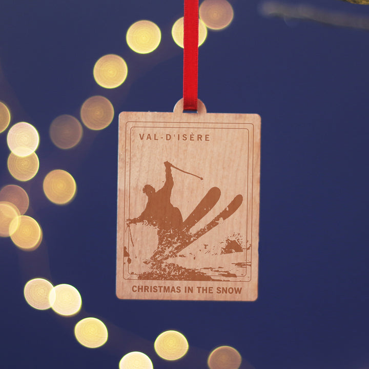 Val-D'Isere travel memory anniversary gift, commemorate your Christmas memories, with this sustainable wood cherry veneered ornament with ta skier. Hanging against a blue background. Personalise the location and text for a perfect letterbox friendly gift for travellers.