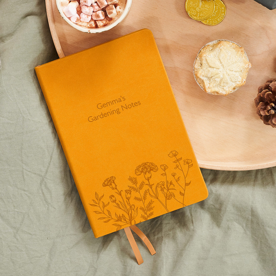 A top down image of a mustard yellow vegan leather notebook. The front of the notebook features a pattern of delicate flowers along the bottom edge. The personalised engraving reads Gemma's Gardening Notes. It is resting on a wooden tray with a mug of hot chocolate, a mince pie, and a pine cone.