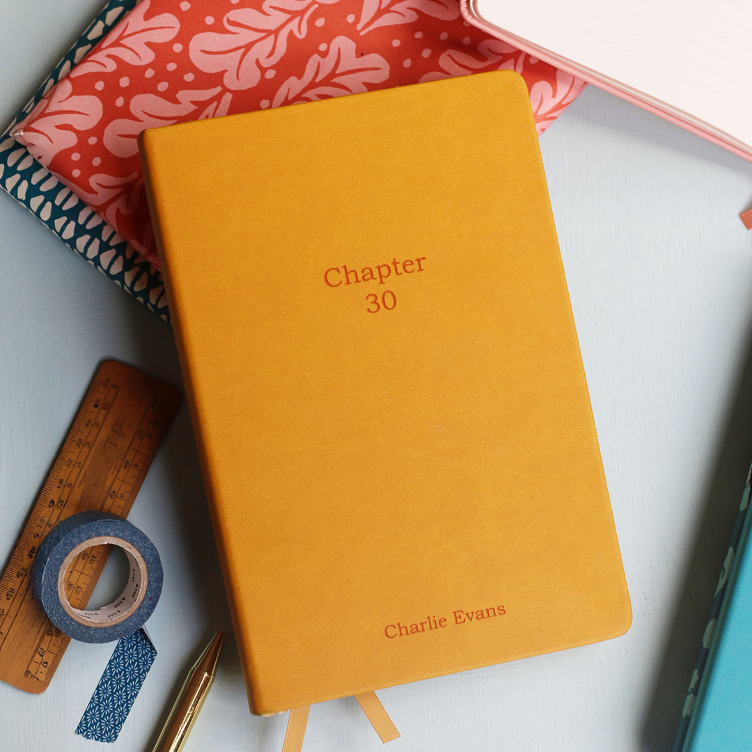 Mustard yellow laser engraved notebook titled 'Chapter 30' with space for a  name for a special 30th birthday present or personalised teacher notebook gift on a white surface with stationery items.