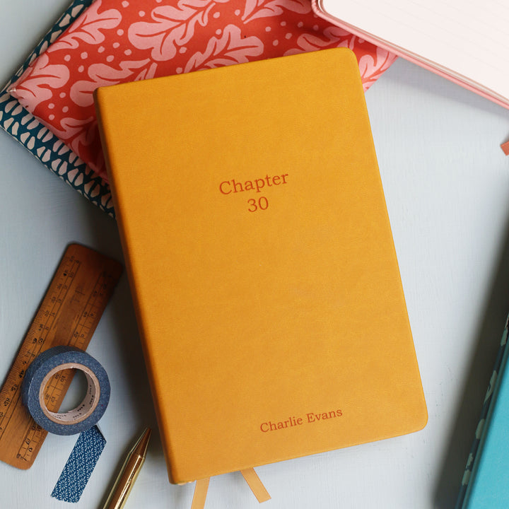 Mustard yellow laser engraved notebook titled 'Chapter 30' with space for a  name for a special 30th birthday present or personalised teacher notebook gift on a white surface with stationery items.