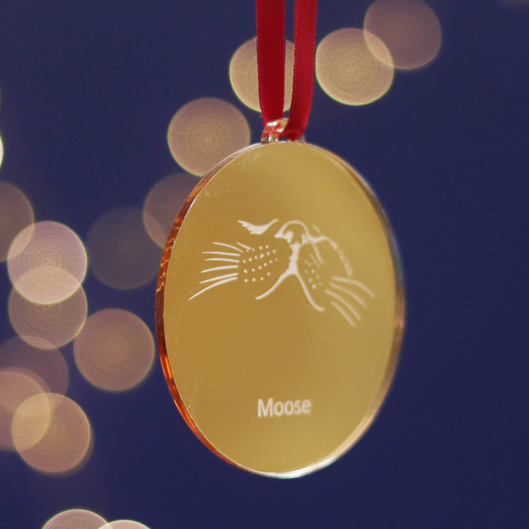 Gold medal-like personalised bauble Christmas tree decoration with 'Moose' engraving on a red ribbon against a blurred light background. For your cats first Christmas or a gift for that number one pet lover in your life.