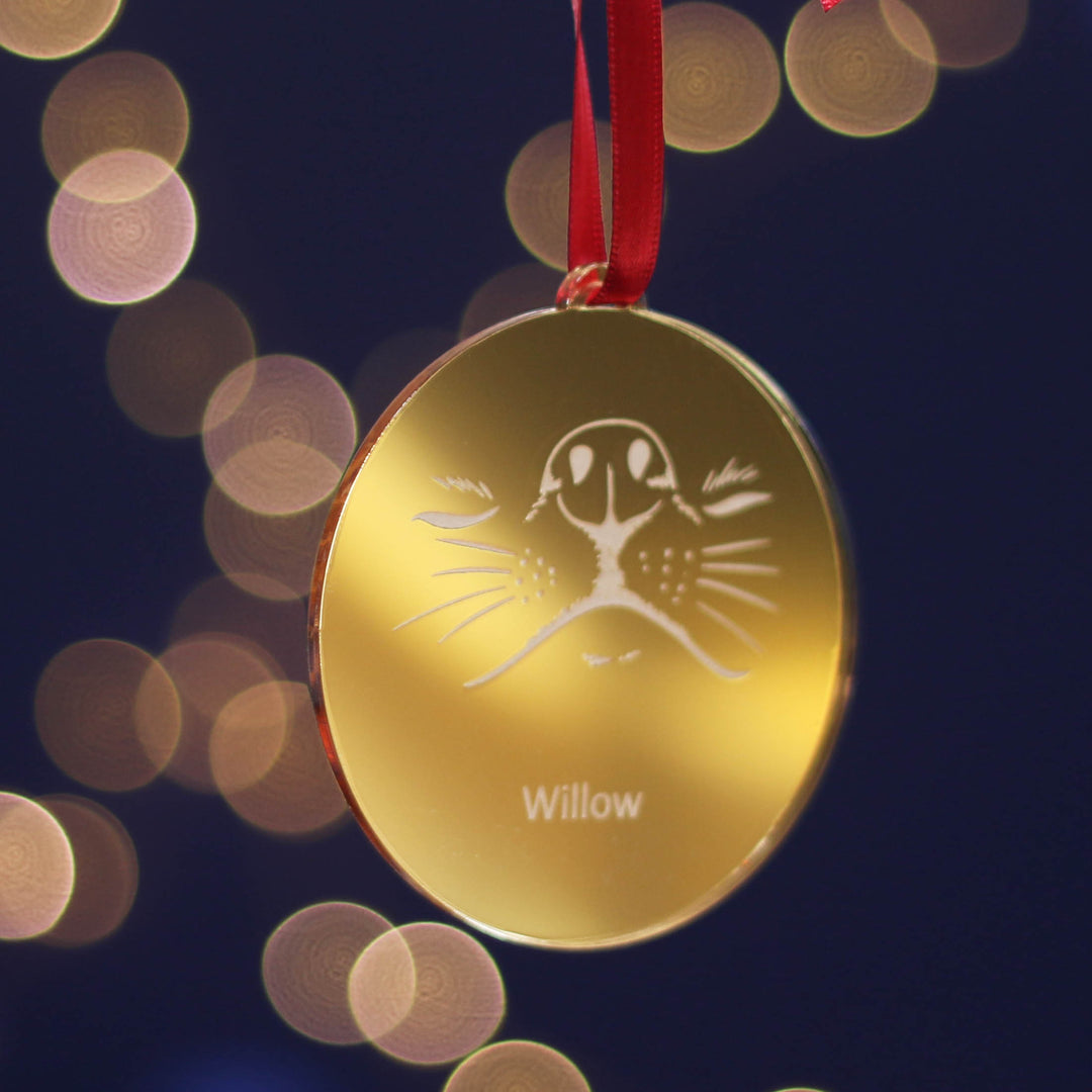 Festive gold personalised Christmas tree bauble decoration ornament with an adorable dog design and custom pet name engraving, hanging on a red ribbon against a dark background with bokeh lights.