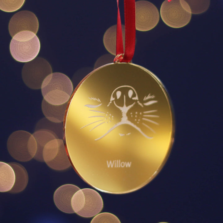 Festive gold personalised Christmas tree bauble decoration ornament with an adorable dog design and custom pet name engraving, hanging on a red ribbon against a dark background with bokeh lights.