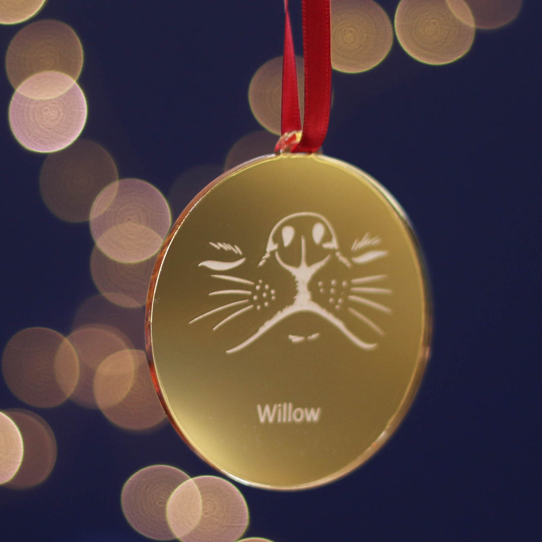 Shining gold special Christmas tree gift decoration ornament with a very cute smushed dog face design and customisable pet name, hanging on red ribbon against a dark background with bokeh lights. A mirrored special Xmas treasure to dazzle this festive season.