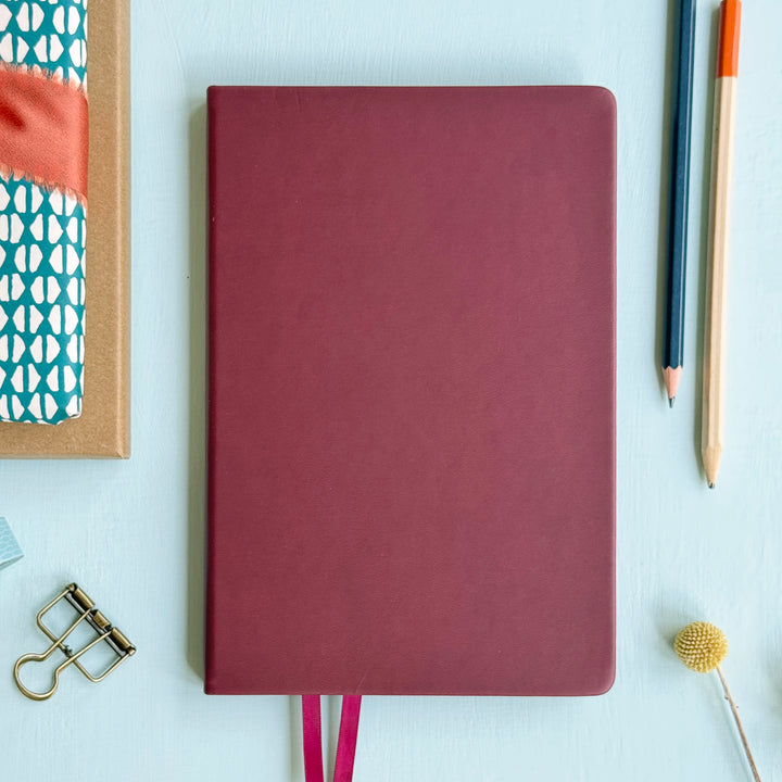 A top down image of a plain simple burgundy pink vegan leather notebook. It is resting on a light blue surface. Surrounding the book are various stationary items.