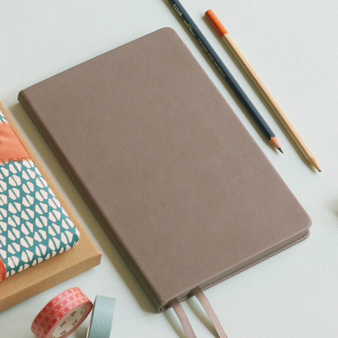 A top down image of a plain simple grey vegan leather notebook. It is resting on a light blue surface. Surrounding the book are various stationary items.