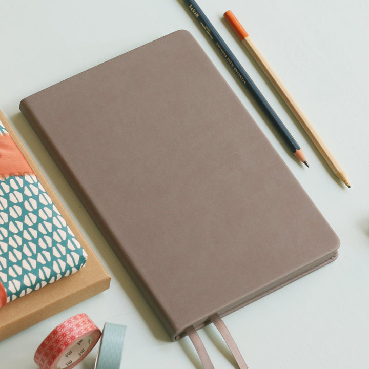 A top down image of a plain simple grey vegan leather notebook. It is resting on a light blue surface. Surrounding the book are various stationary items.
