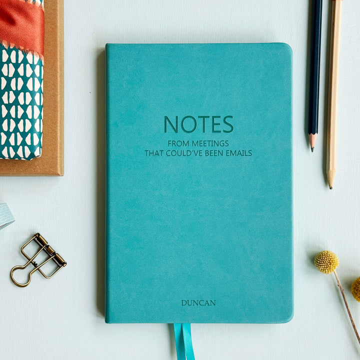 Turquoise personalised notebook with 'NOTES FROM MEETINGS THAT COULD'VE BEEN EMAILS' text and placed on a white surface with office supplies.