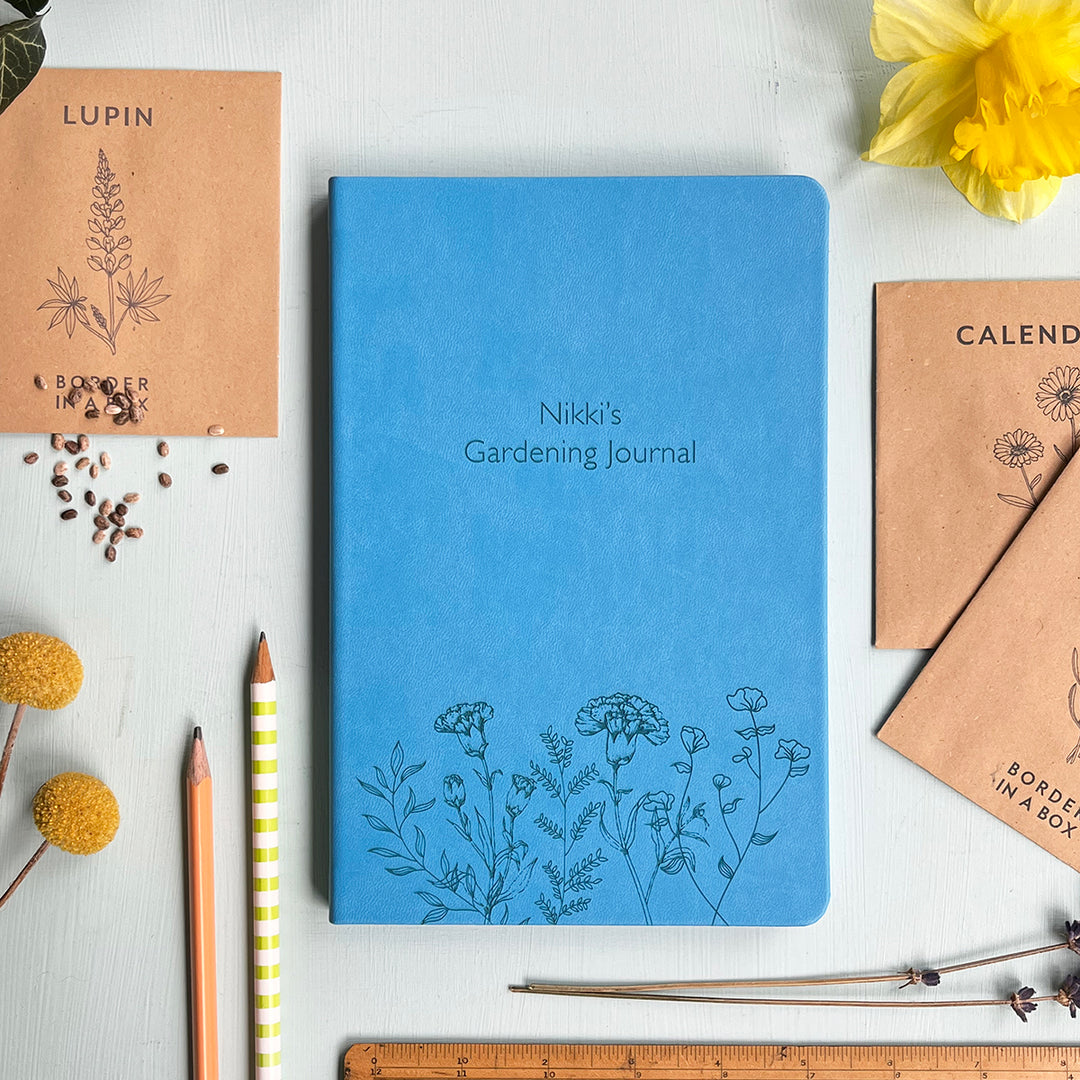 A top down image of a blue vegan leather notebook resting on a pale aqua table. The front of the notebook features a pattern of delicate flowers along the bottom edge. The personalised engraving reads Nikkis Gardening Journal. Surrounding the book are various stationary items.
