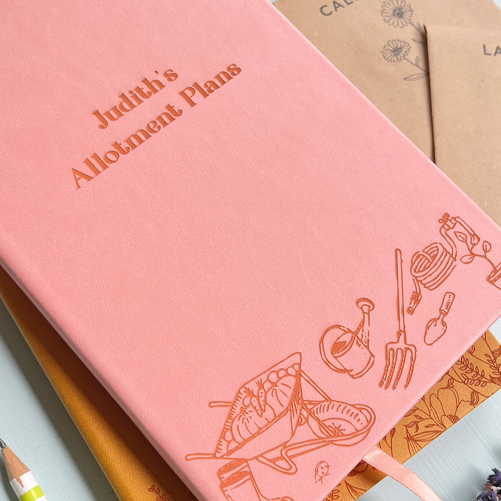 A close up  image of a pink vegan leather notebook resting on a pale aqua table. The front of the notebook features an engraving of gardening tools along the bottom edge. The personalised text reads "Judith's Allotment Plans". Surrounding the book are flower seed packets.