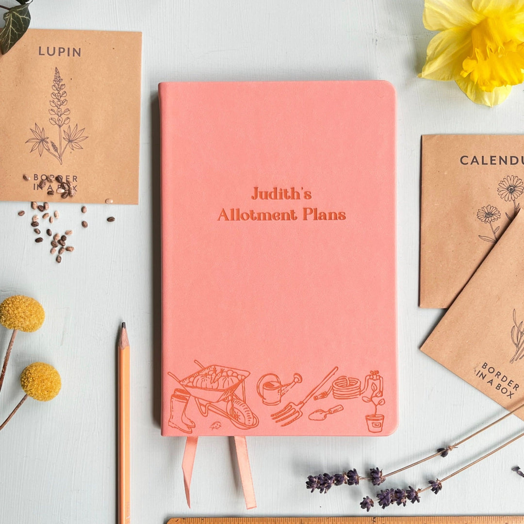 A top down image of a pink vegan leather notebook resting on a pale aqua table. The front of the notebook features an engraving of gardening tools along the bottom edge. The personalised text reads "Judith's Allotment Plans". Surrounding the book are various flower seed packets and dried flowers.