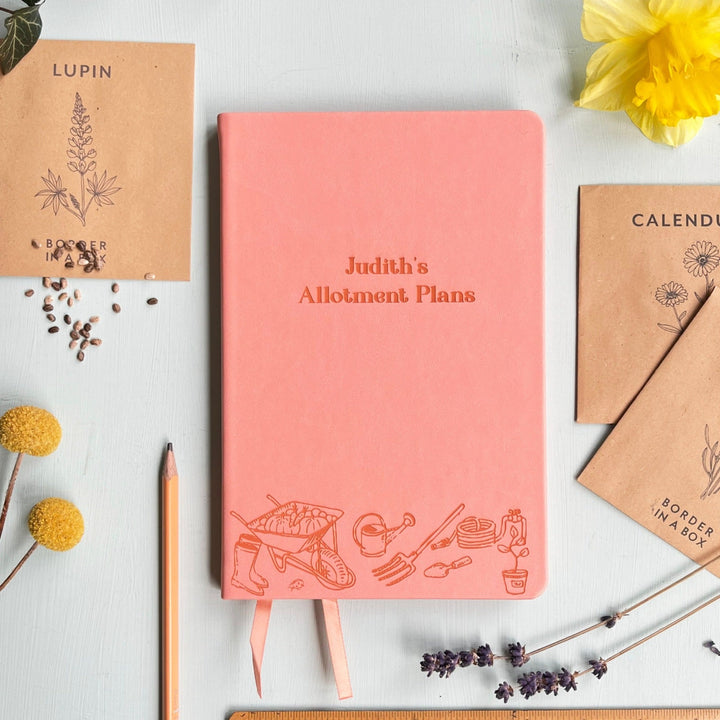 A top down image of a pink vegan leather notebook resting on a pale aqua table. The front of the notebook features an engraving of gardening tools along the bottom edge. The personalised text reads "Judith's Allotment Plans". Surrounding the book are various flower seed packets and dried flowers.
