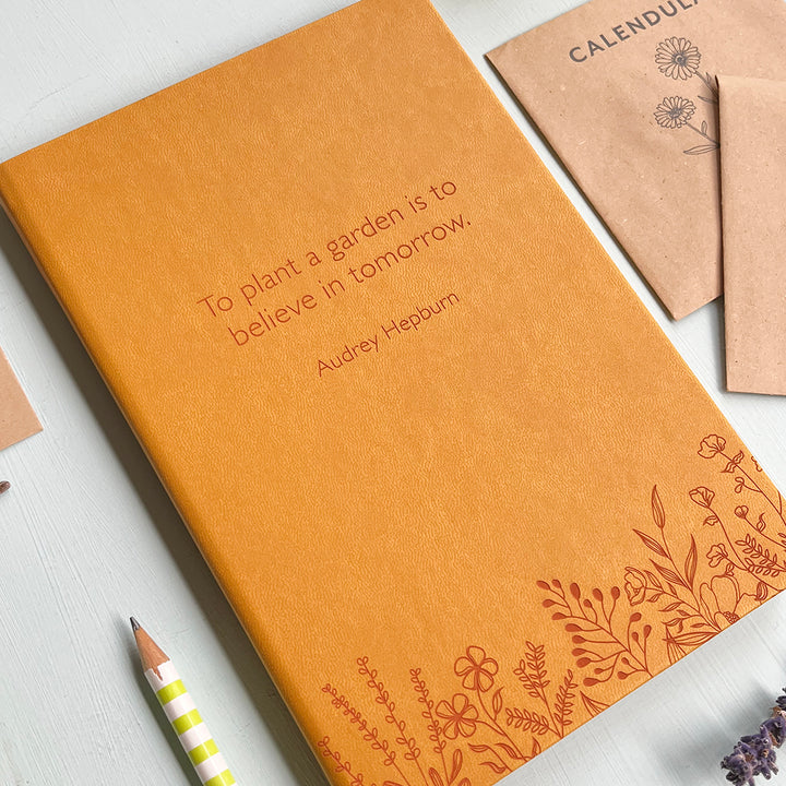 A mustard yellow vegan leather notebook resting on a pale aqua table. The front of the notebook features a pattern of delicate flowers along the bottom edge. The personalised engraving reads "To plant a garden is to believe in tomorrow. - Audrey Hepburn". Surrounding the book are various stationary items and dried flowers.