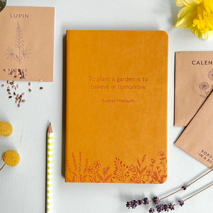 A top down image of a yellow vegan leather notebook resting on a pale aqua table. The front of the notebook features a pattern of delicate flowers along the bottom edge. The personalised engraving reads "To plant a garden is to believe in tomorrow. - Audrey Hepburn". Surrounding the book are various stationary items and dried flowers and seeds.
