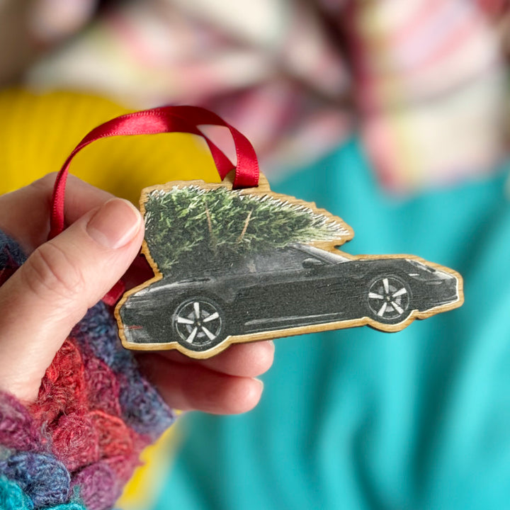 A hand holding a wooden car enthusiasts gift Porsche 911 Christmas tree decoration with a black car and green tree on a blue and yellow background.